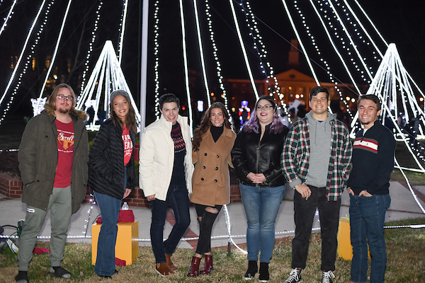A group of 7 students attending Lighting the Quad and standing in front of a decoration of lights in the shape of a tree. 