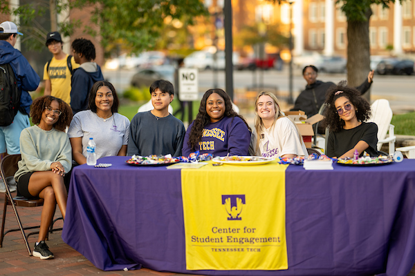 A group of students sitting at a Student Engagement information table smiling at the camera.