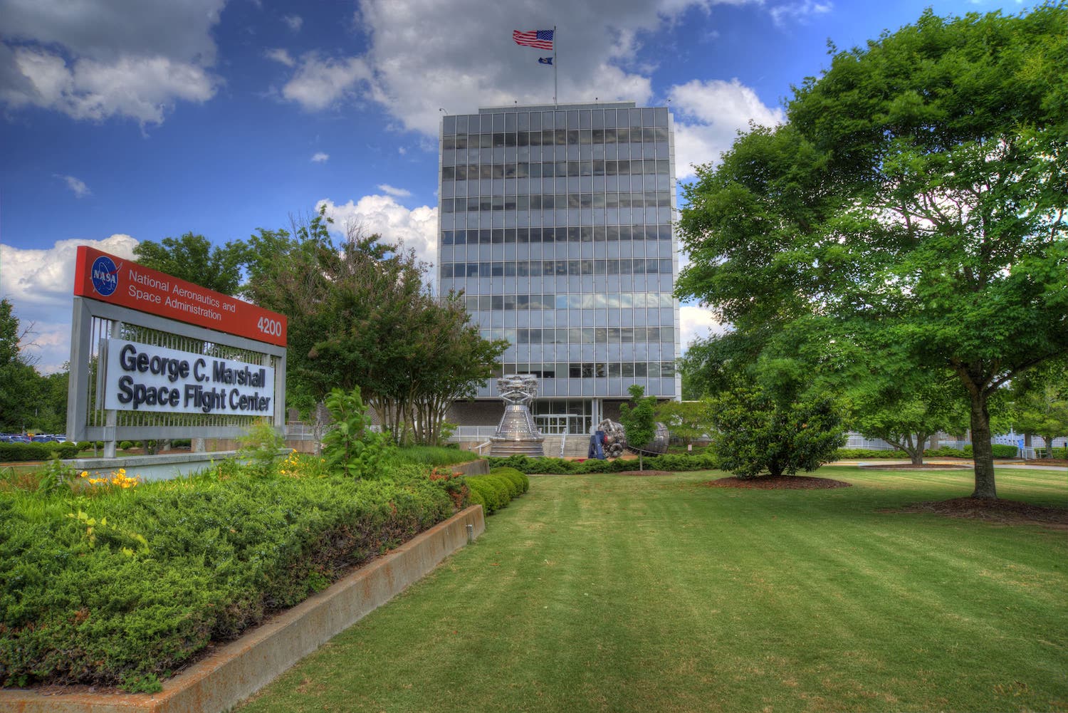 A beautiful, sunny day with a blue sky and white clouds. There is a mowed lawn with a tall building; an American Flag on top; a sign reading George C. Marshall Space Flight Center 