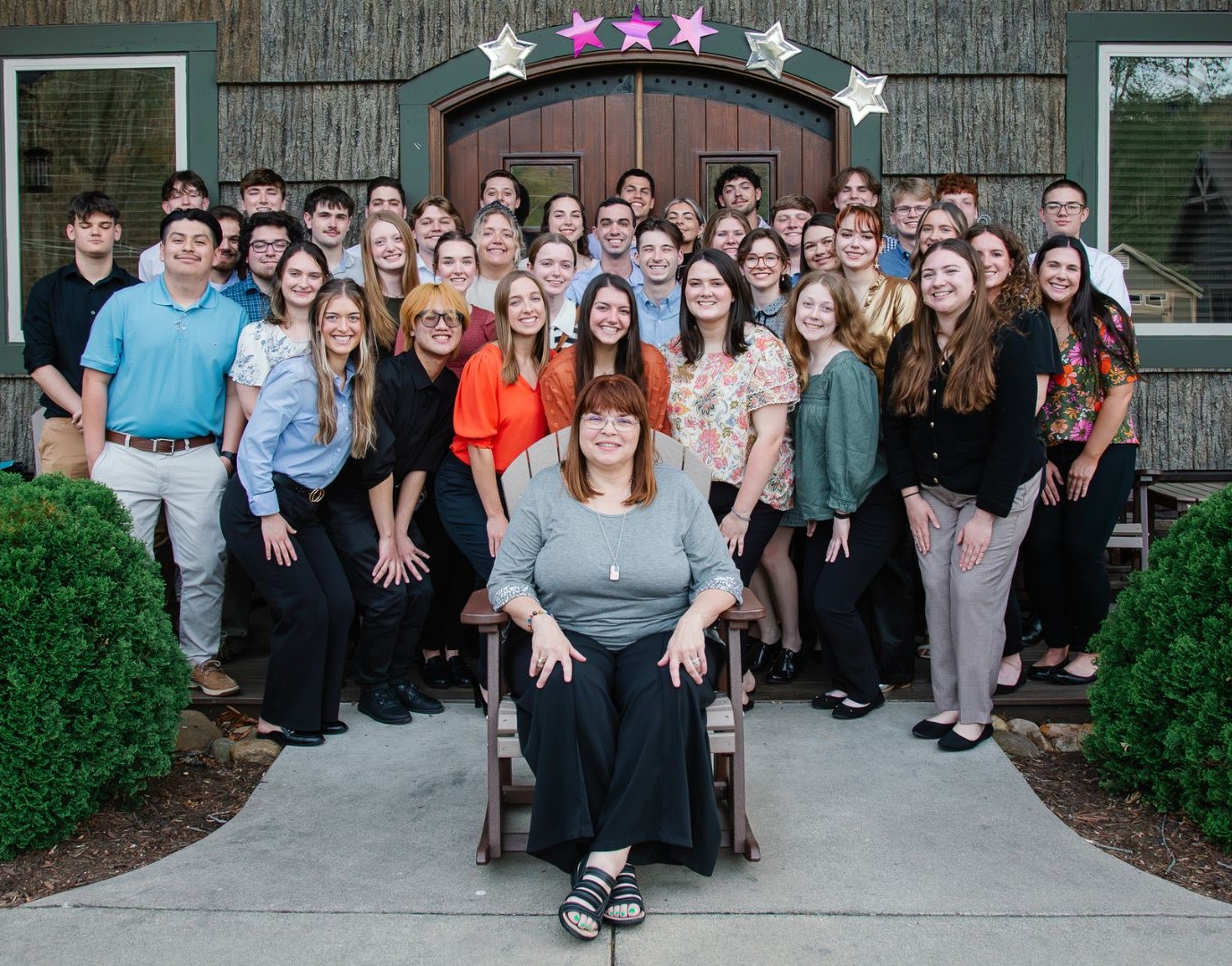 Large group of students pose outdoors in front of a building, with Mrs. Sherrie Cannon seated at the center and smiling at the camera.