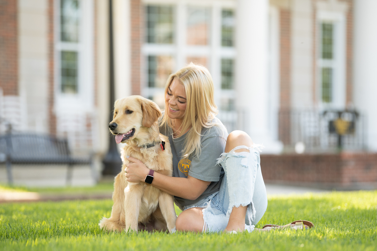 Female student sits in the grass petting a golden retriever.