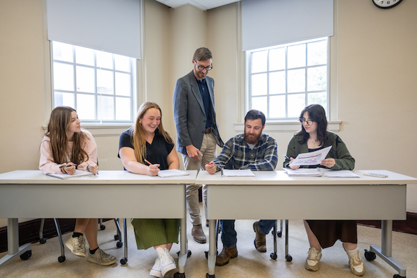 An instructor assists four students reviewing worksheets together at classroom tables.