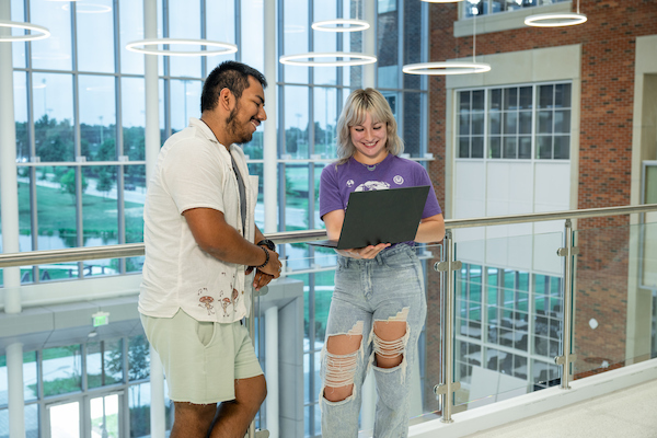 Two students smile while looking at a laptop together beside a glass railing in a bright campus atrium.