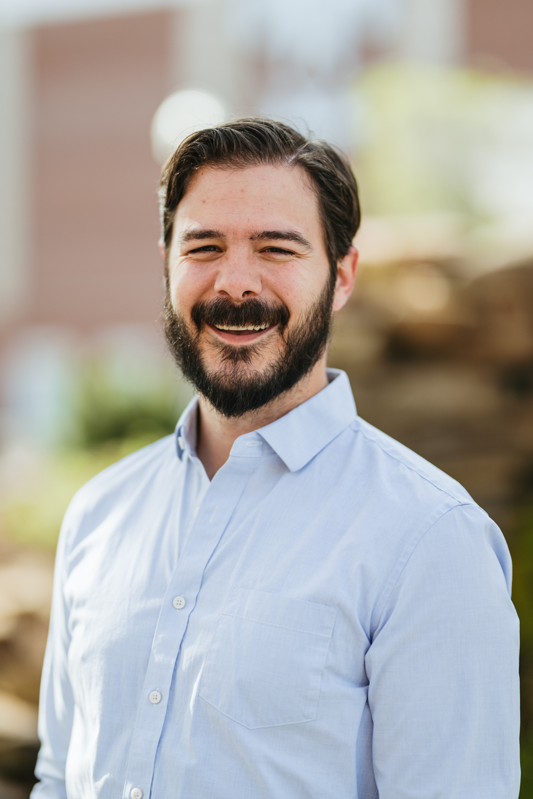 A male with dark brown hair and dark brown beard wearing a button up shirt.