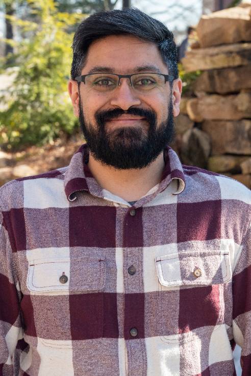 Asian Indian mail wearing glasses, with a beard, wearing a maroon and white plaid checkered button up shirt.