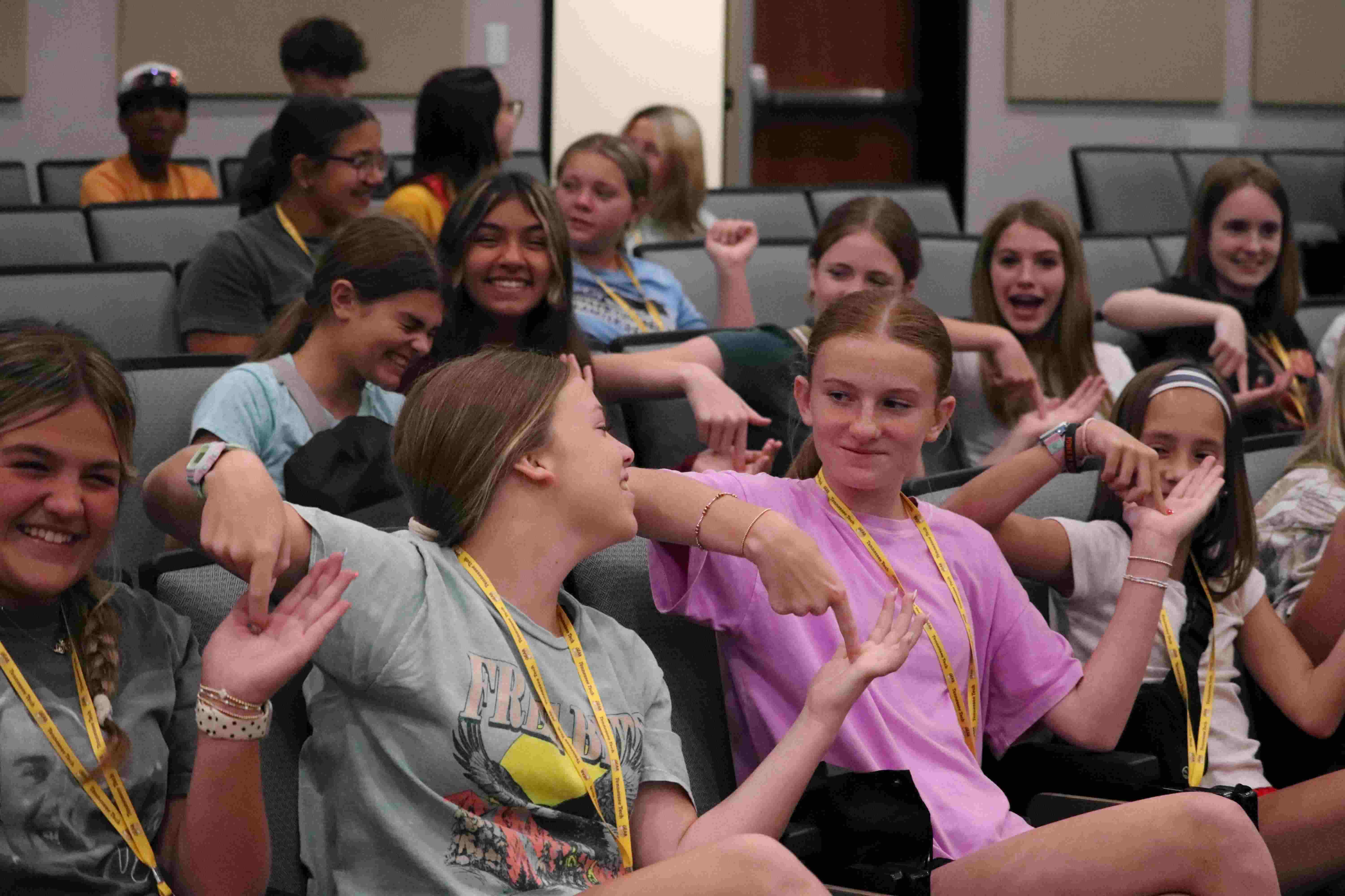 A group of cheerful young students sitting in stadium-style auditorium seating, smiling, chatting, and playfully pointing at each other.