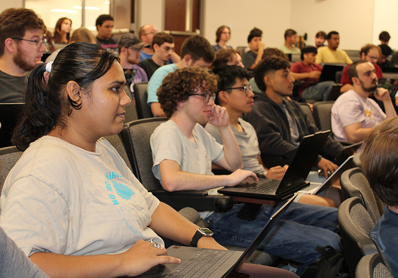 Group of students sitting in an auditorium with their laptops as they listen to a speaker