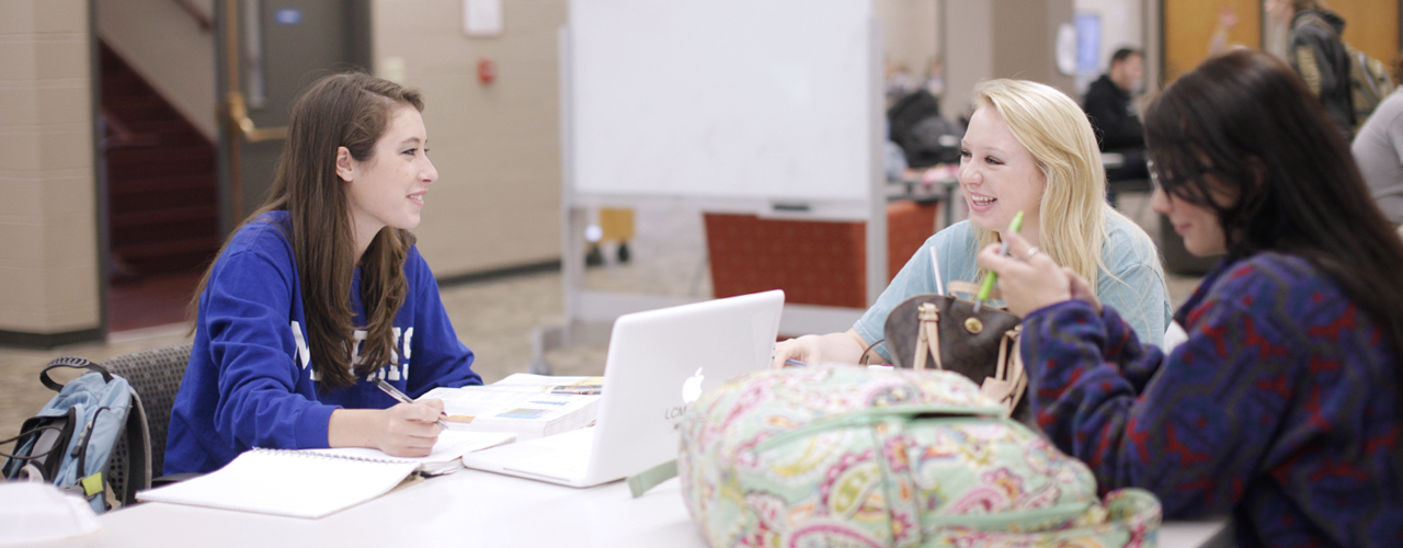 three females studying with laptops, backpacks, and a white board