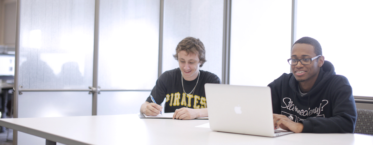 Two male students study with a laptop and notebook at a library table.
