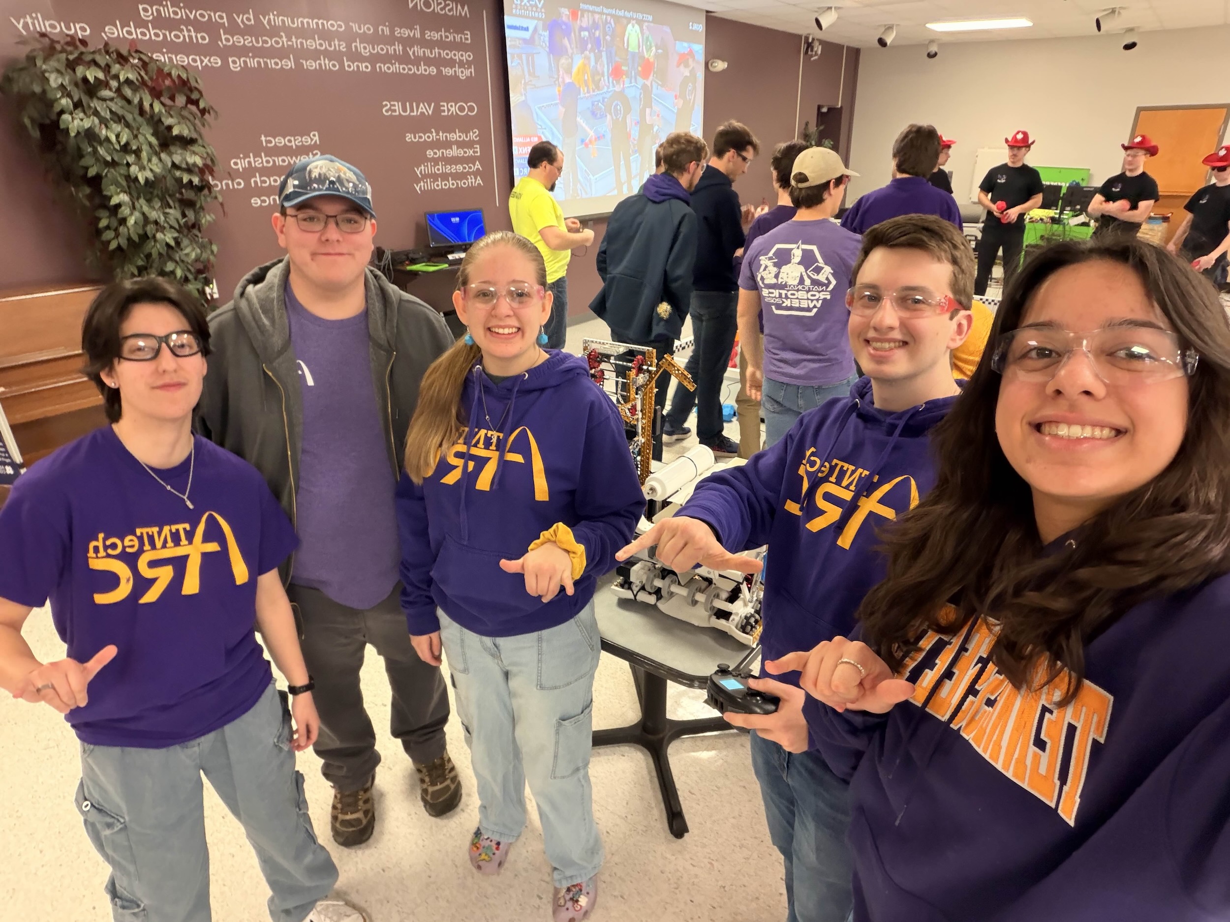 Students in purple and gold attire flash a 'wings up' signal. Others are seen in the background.