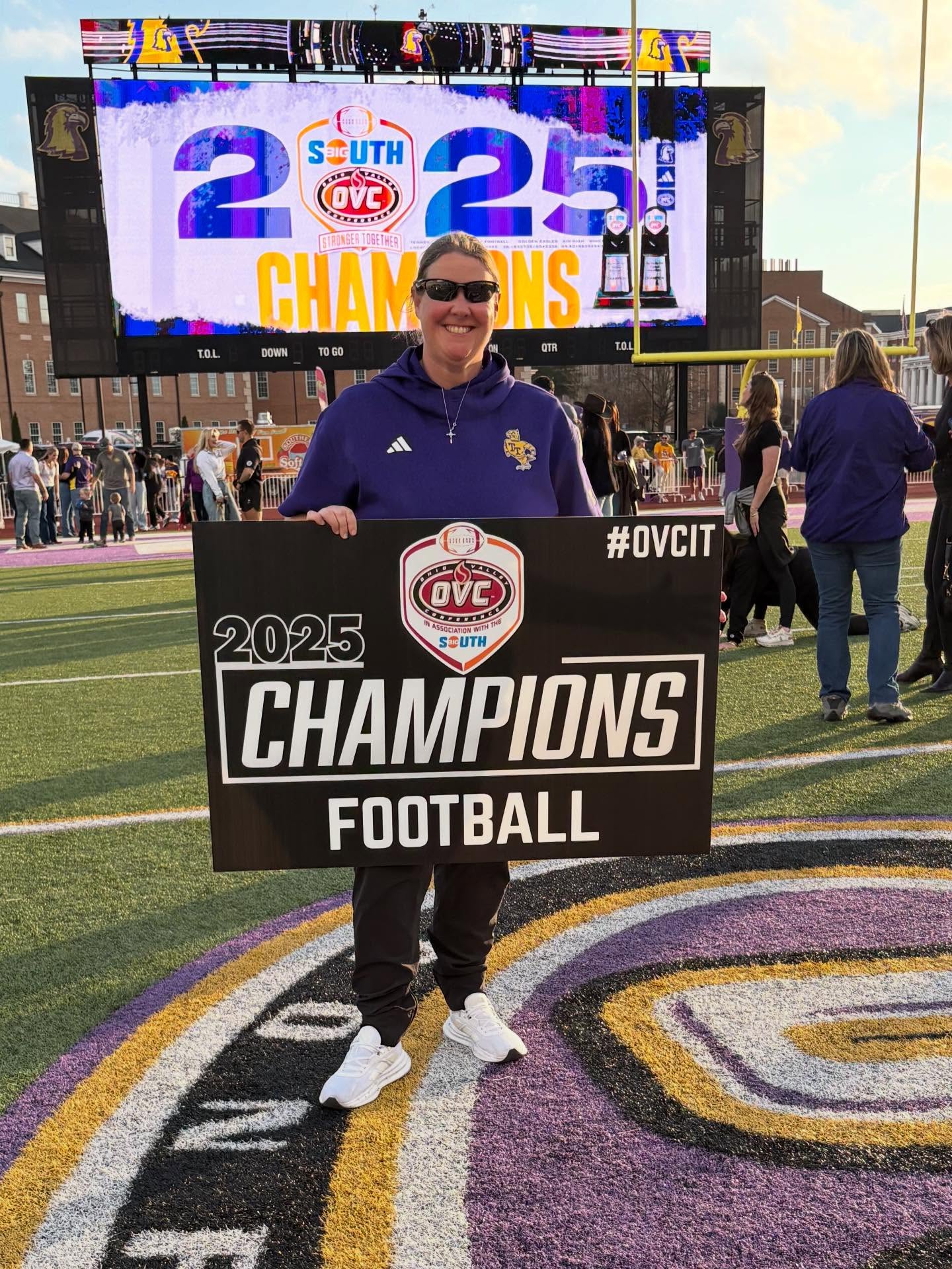 Jennifer Winningham stands on the football field in sunglasses holding an 'OVC Champions' sign.