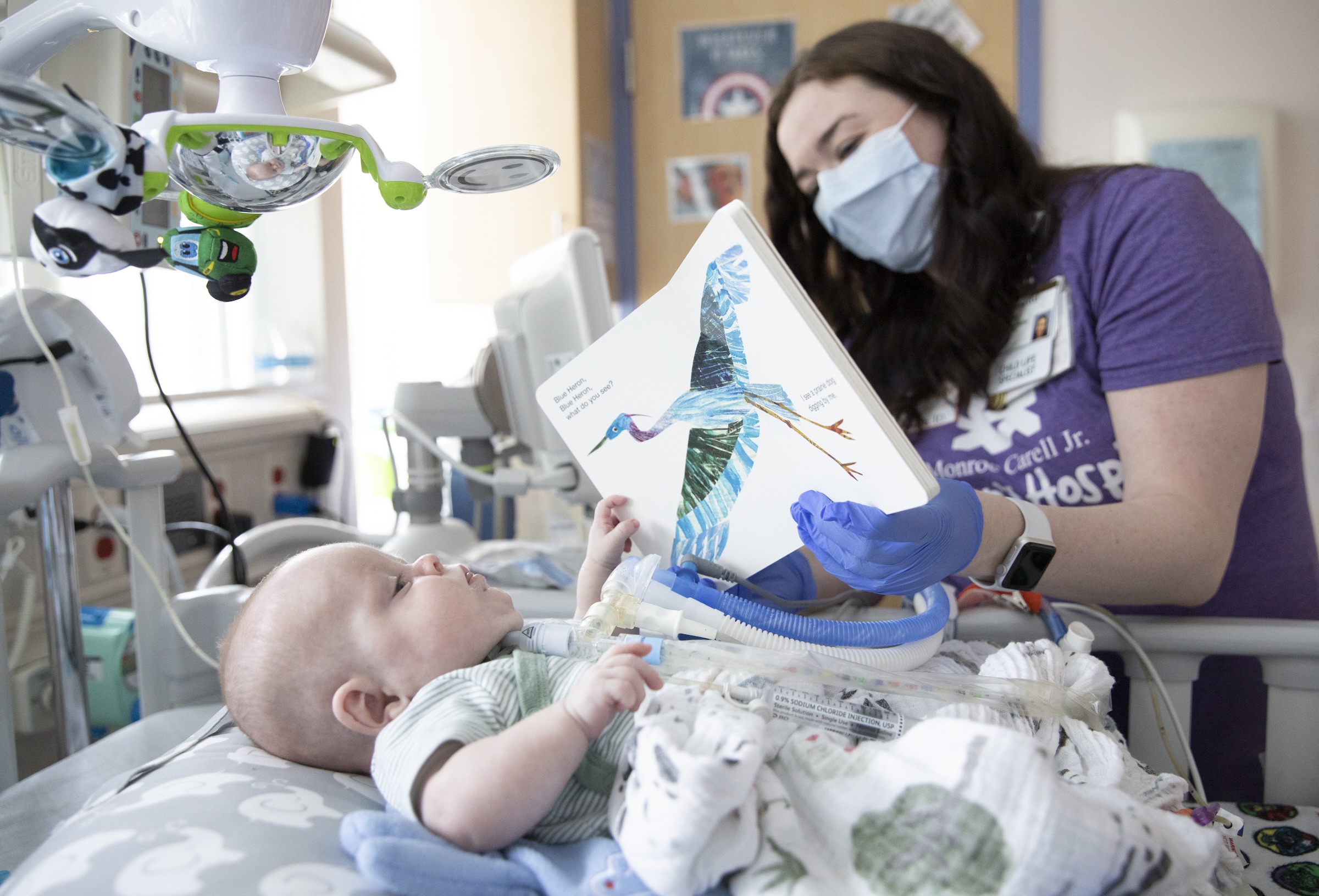 Katy Hoskins wears a mask and holds up a book to a baby who points at a picture. 