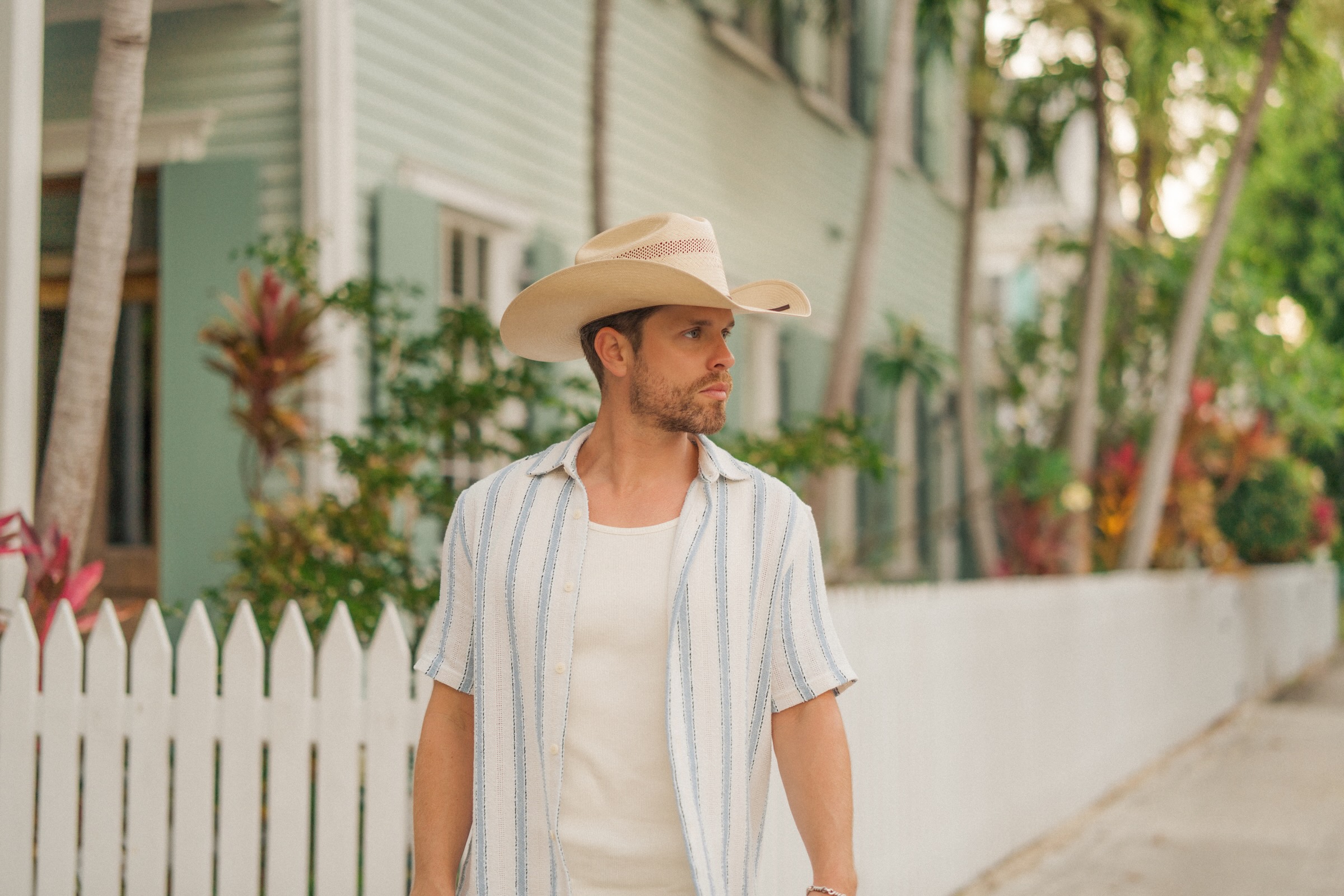 Dustin Lynch is pictured outdoors in a cowboy hat standing in front of a white fence. 