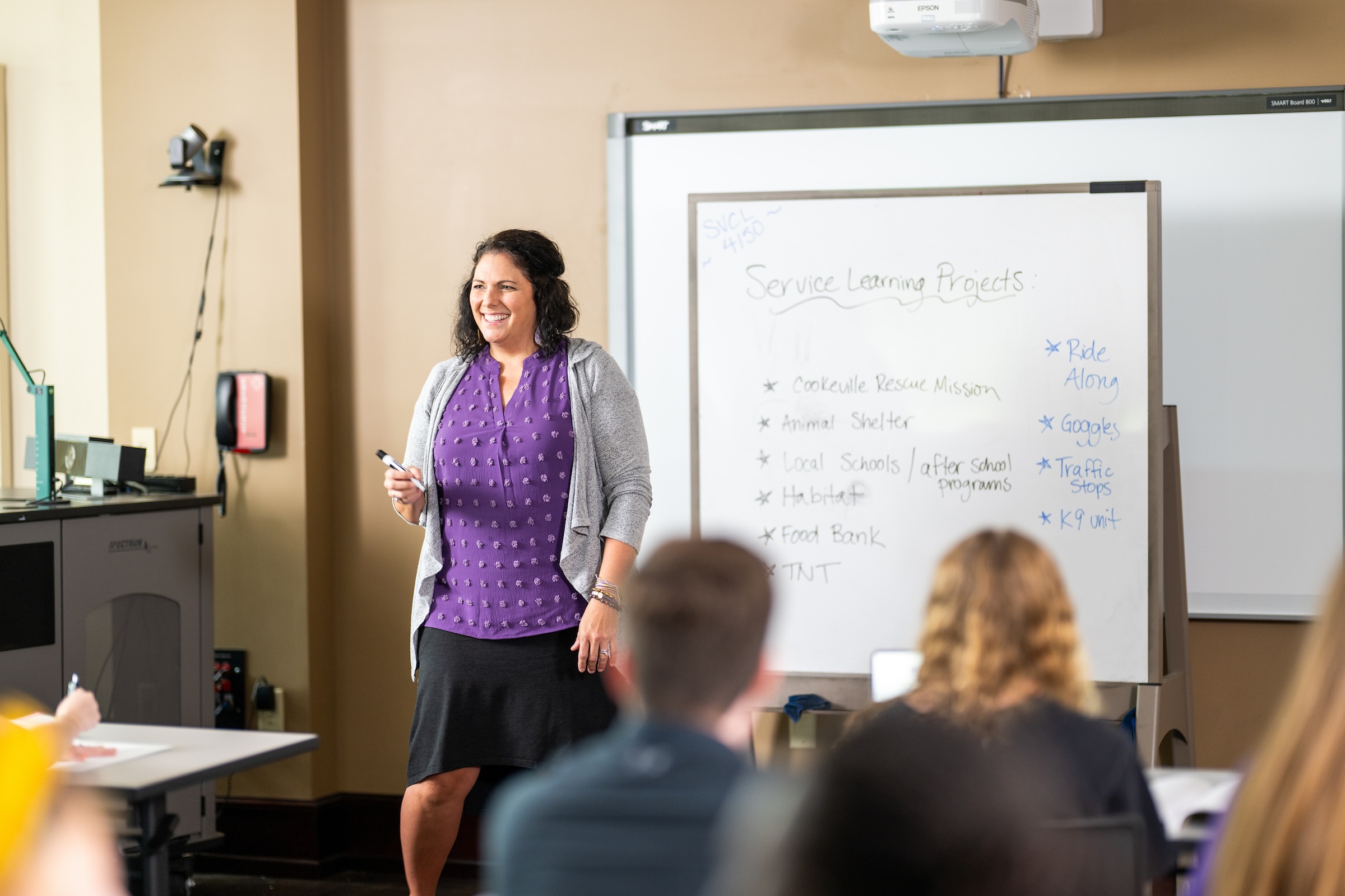 Julie Baker stands in front of a white board and smiles as students look on. 