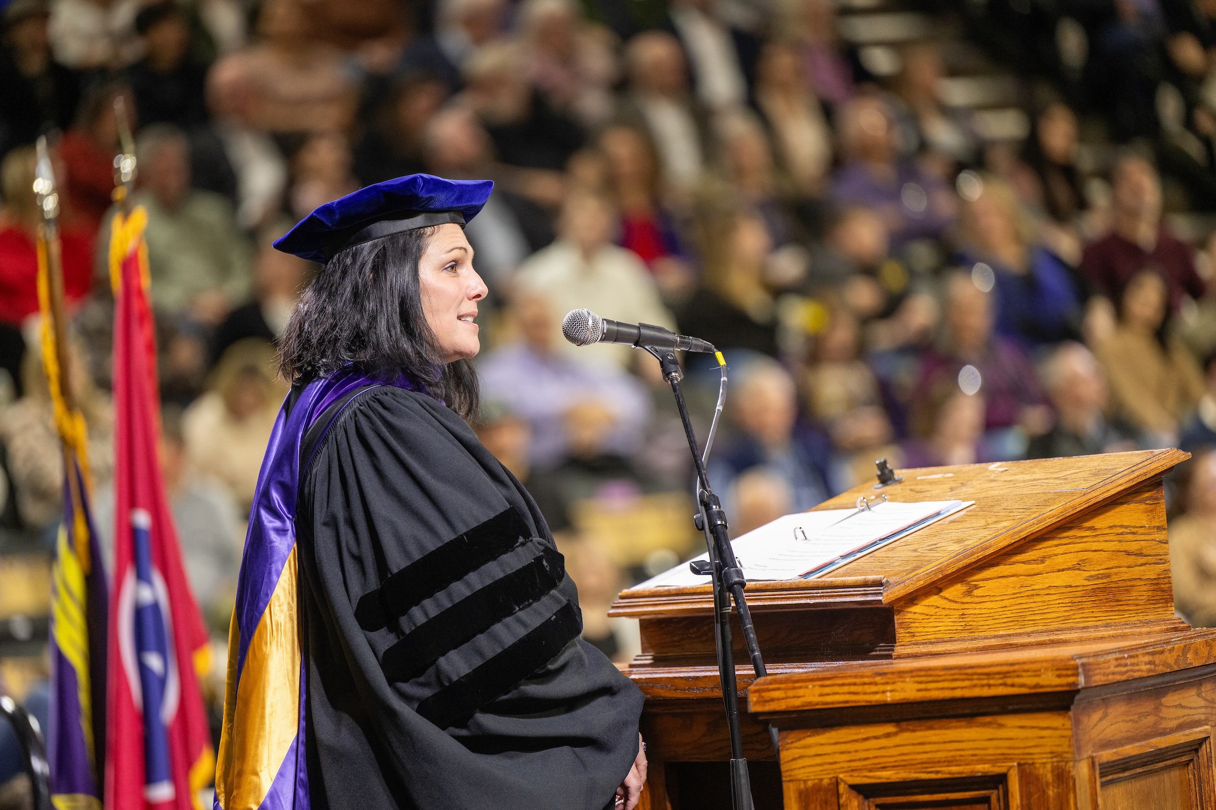 Julie Baker stands at a lectern in academic regalia and smiles as a large audience looks on. 