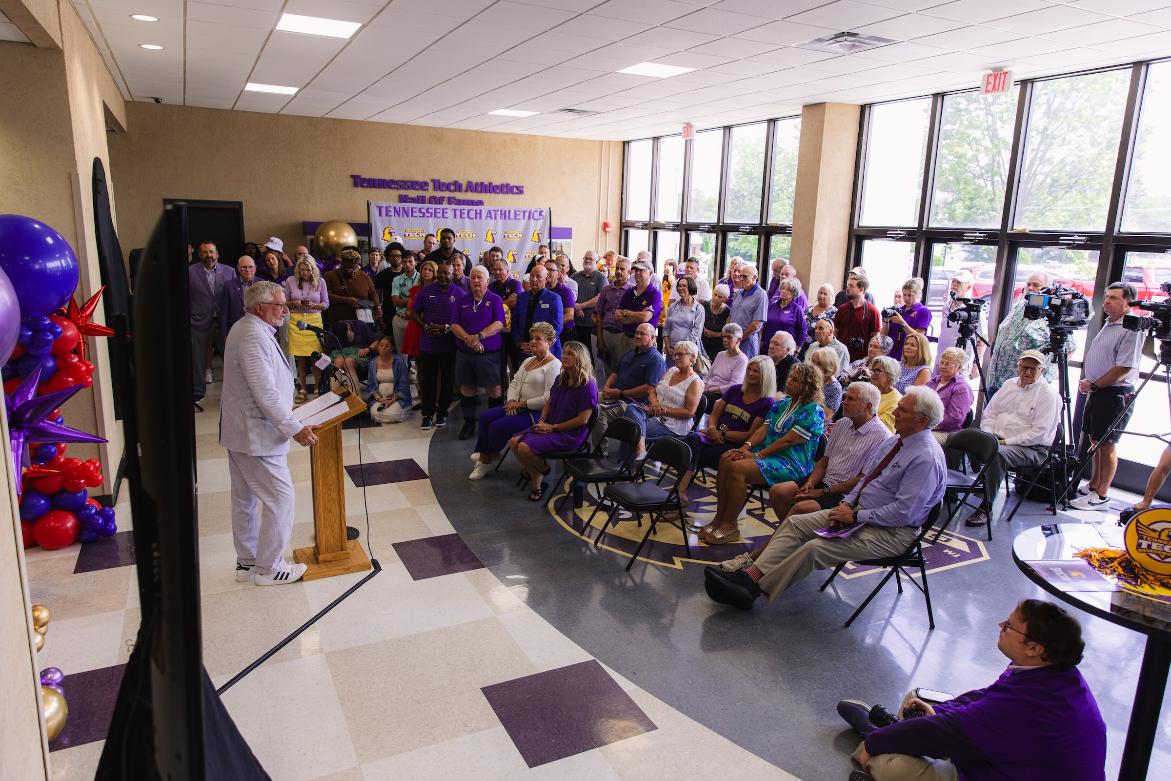President Oldham stands at a podium before a crowded room of fans clad in mostly purple attire. 