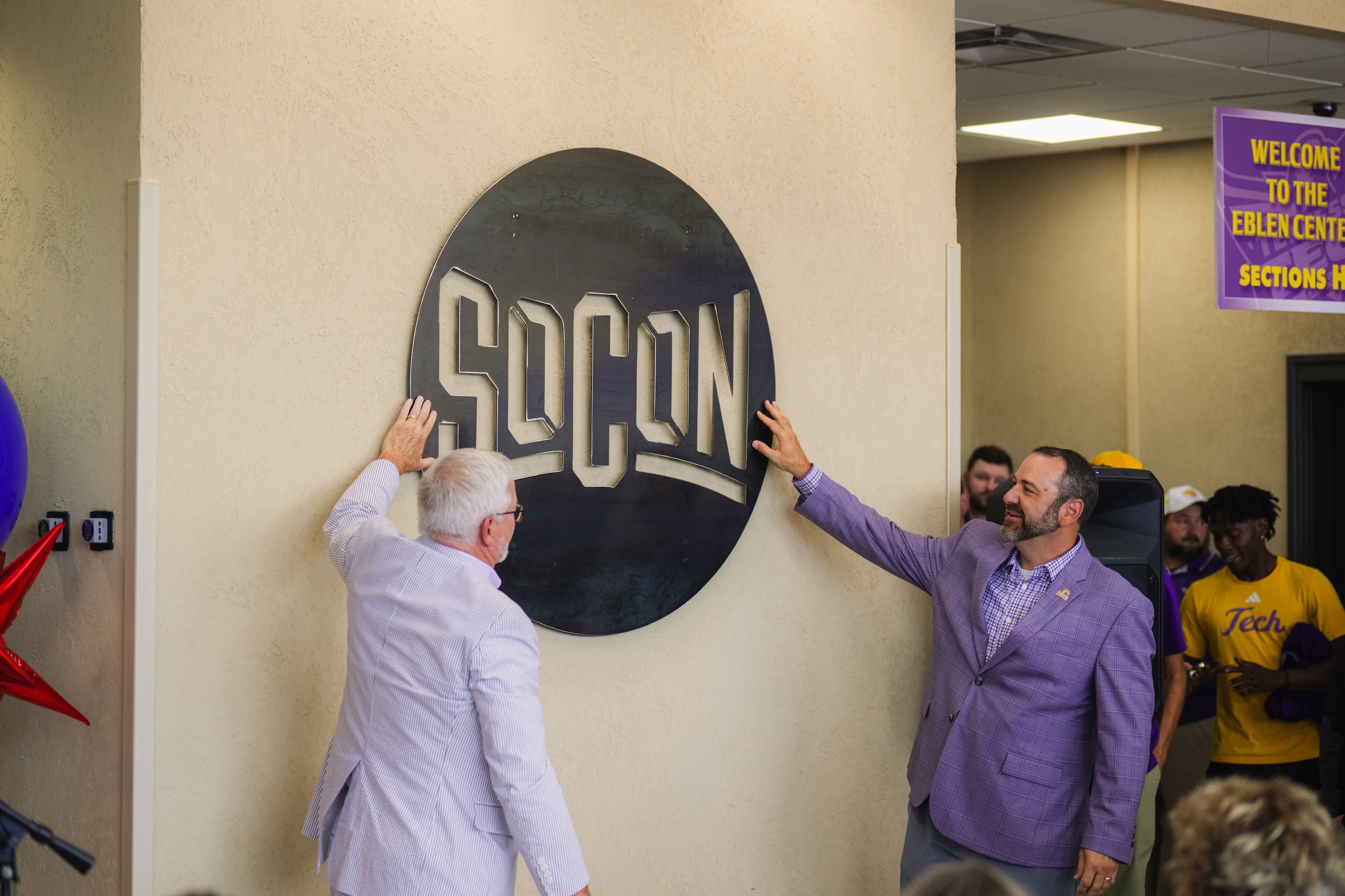 Two men stand at a wall to unveil a new logo that says "SoCon" in a circular frame. 