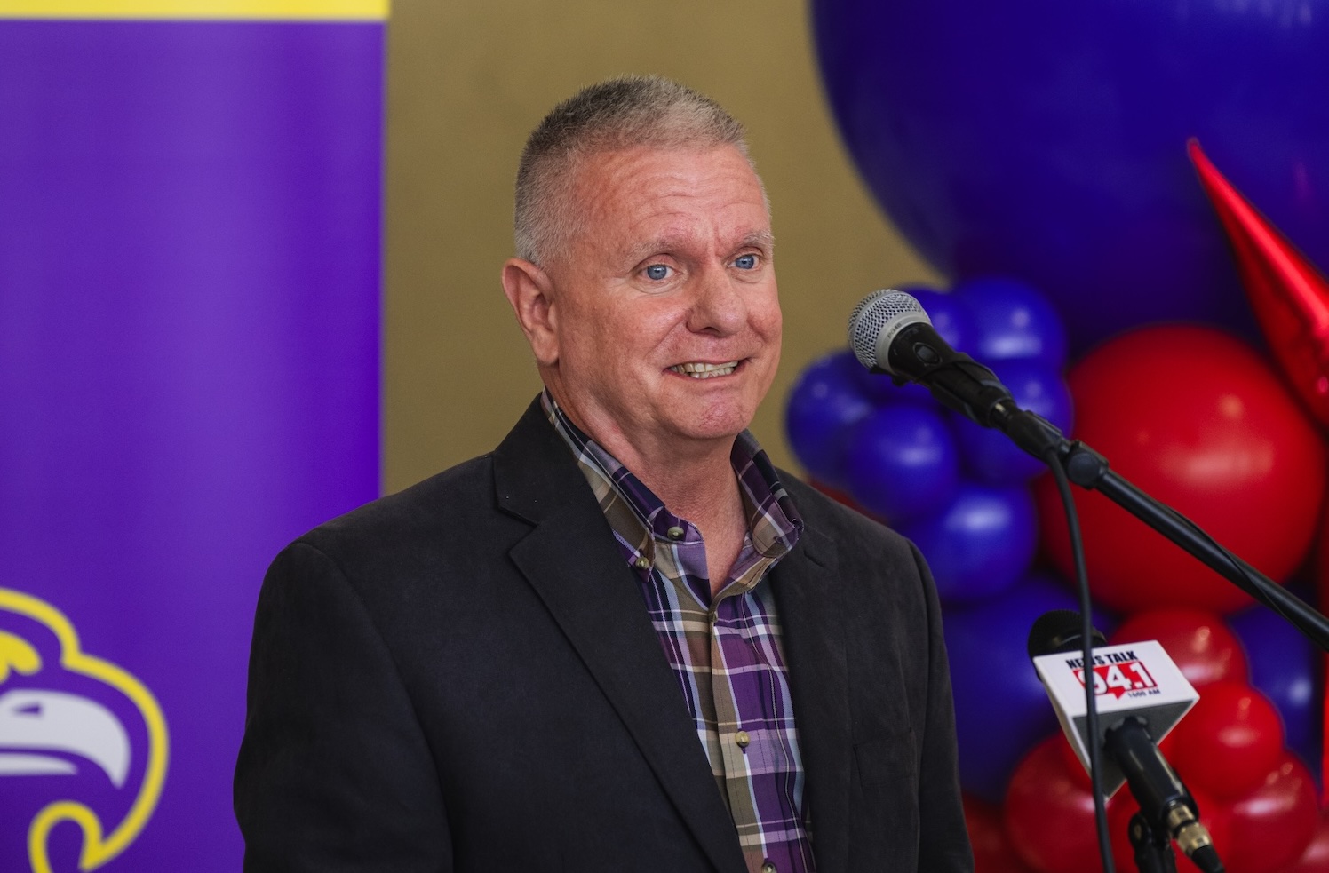Geoff Cabe stands at a podium in front of a balloon display and a purple banner. 
