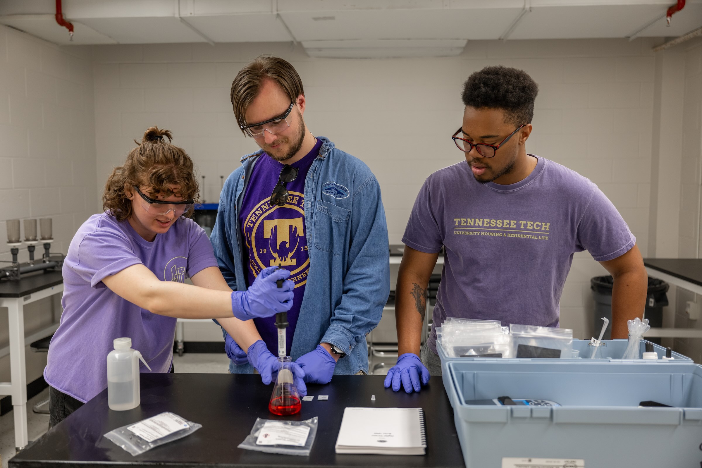 Three students are shown standing over a measuring beaker in a lab space. 