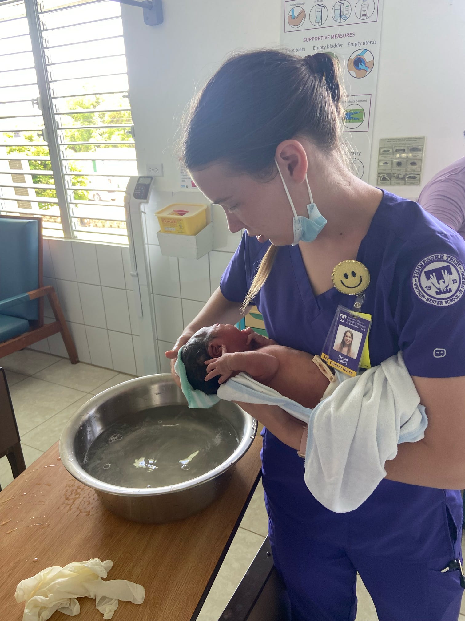 A woman in nurse's scrubs holds a newborn baby next to a water basin. 