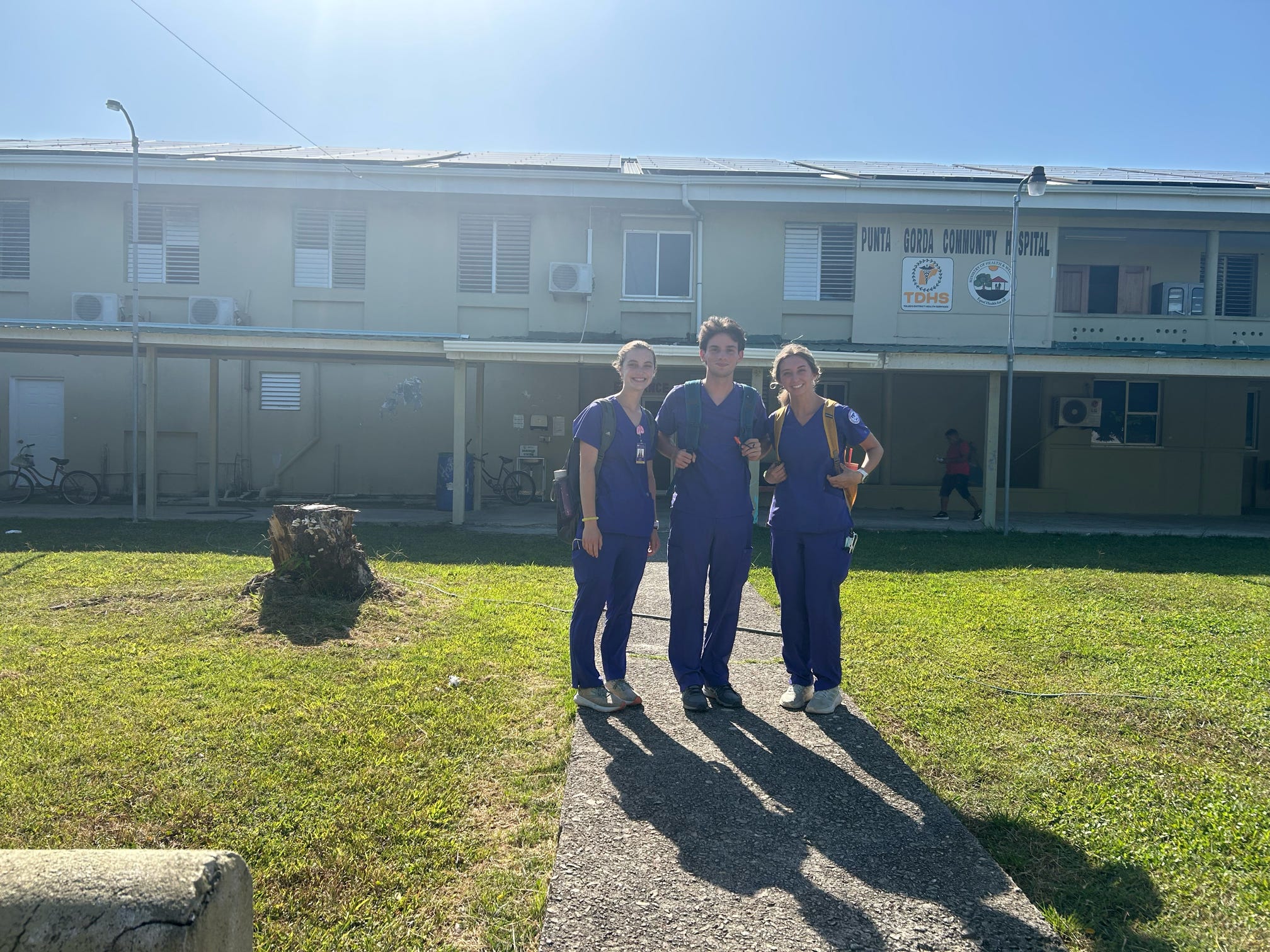 Three individuals in scrubs stand in front of a rustic looking hospital building. 