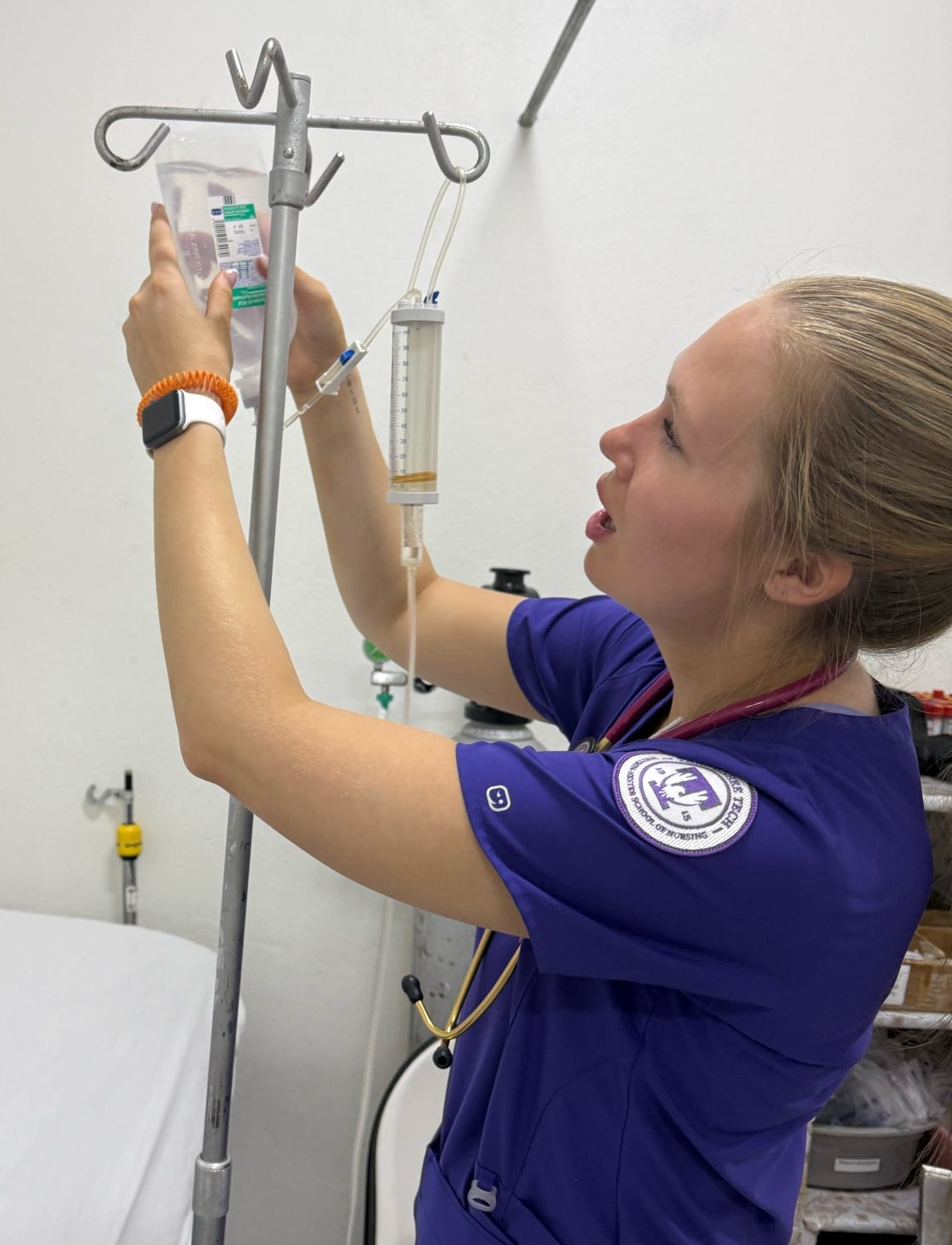 A student in nurse's scrubs adjusts a medication bag on an IV cart.