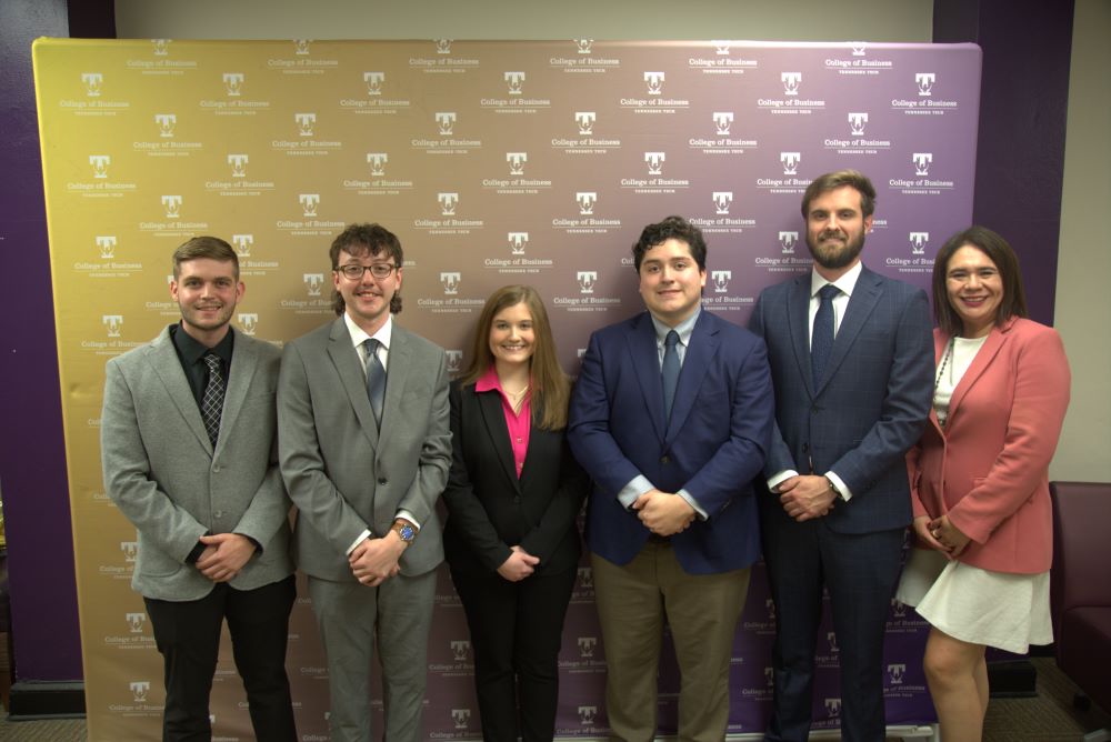 Five students and a professor stand in front of a step-and-repeat and smile. 