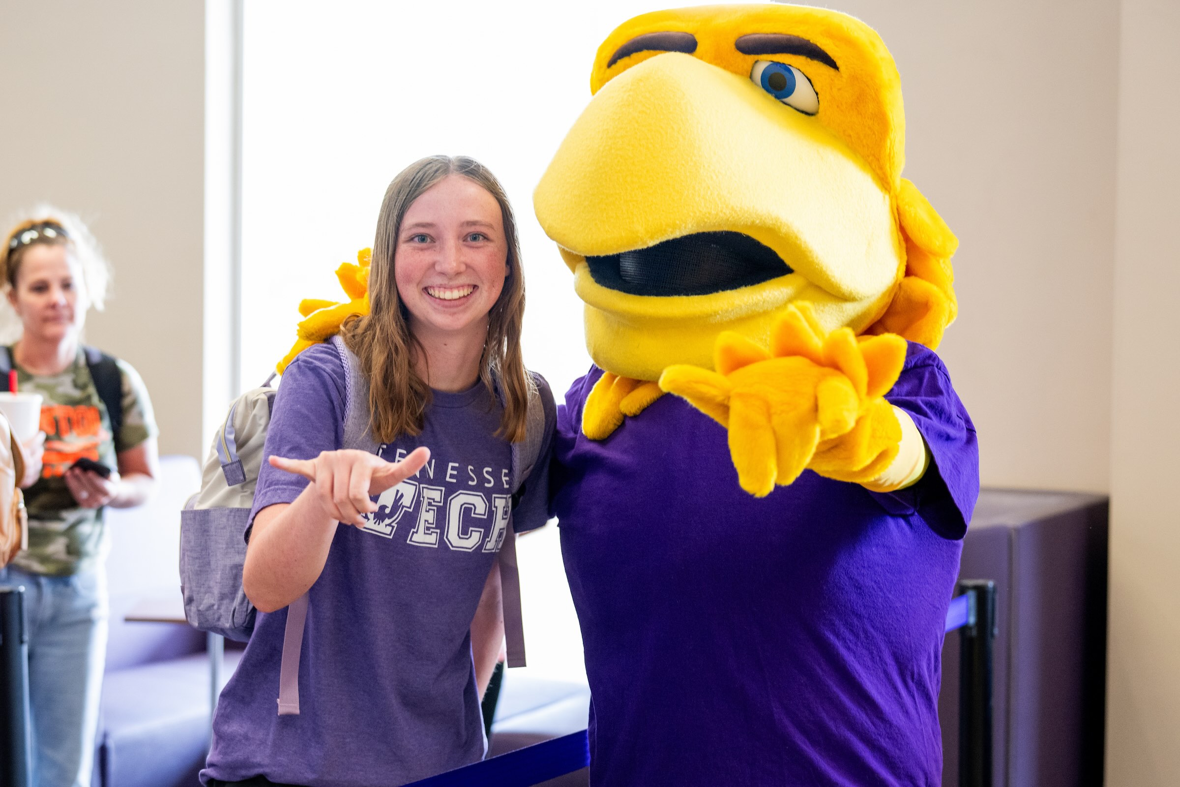 A student in a purple Tech shirt stands next to Awesome Eagle, smiles and gives a 'Wings Up'