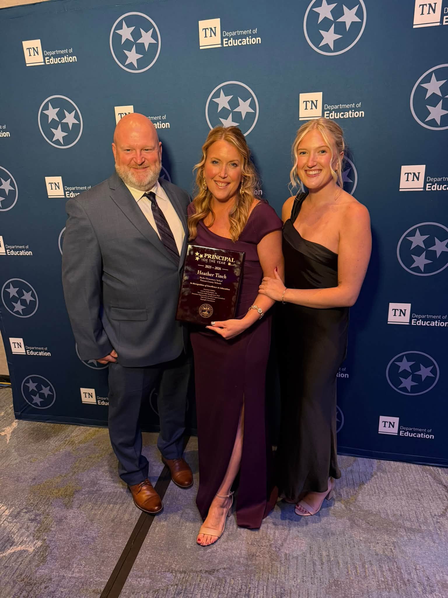 Heather Tinch stands with her husband and daughter in formal attire holding an award placard.