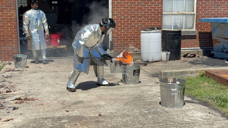 A man in protective gear handles molten aluminum with a large flame. 