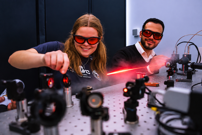 Two individuals in special safety glasses work with quantum equipment and smile. 