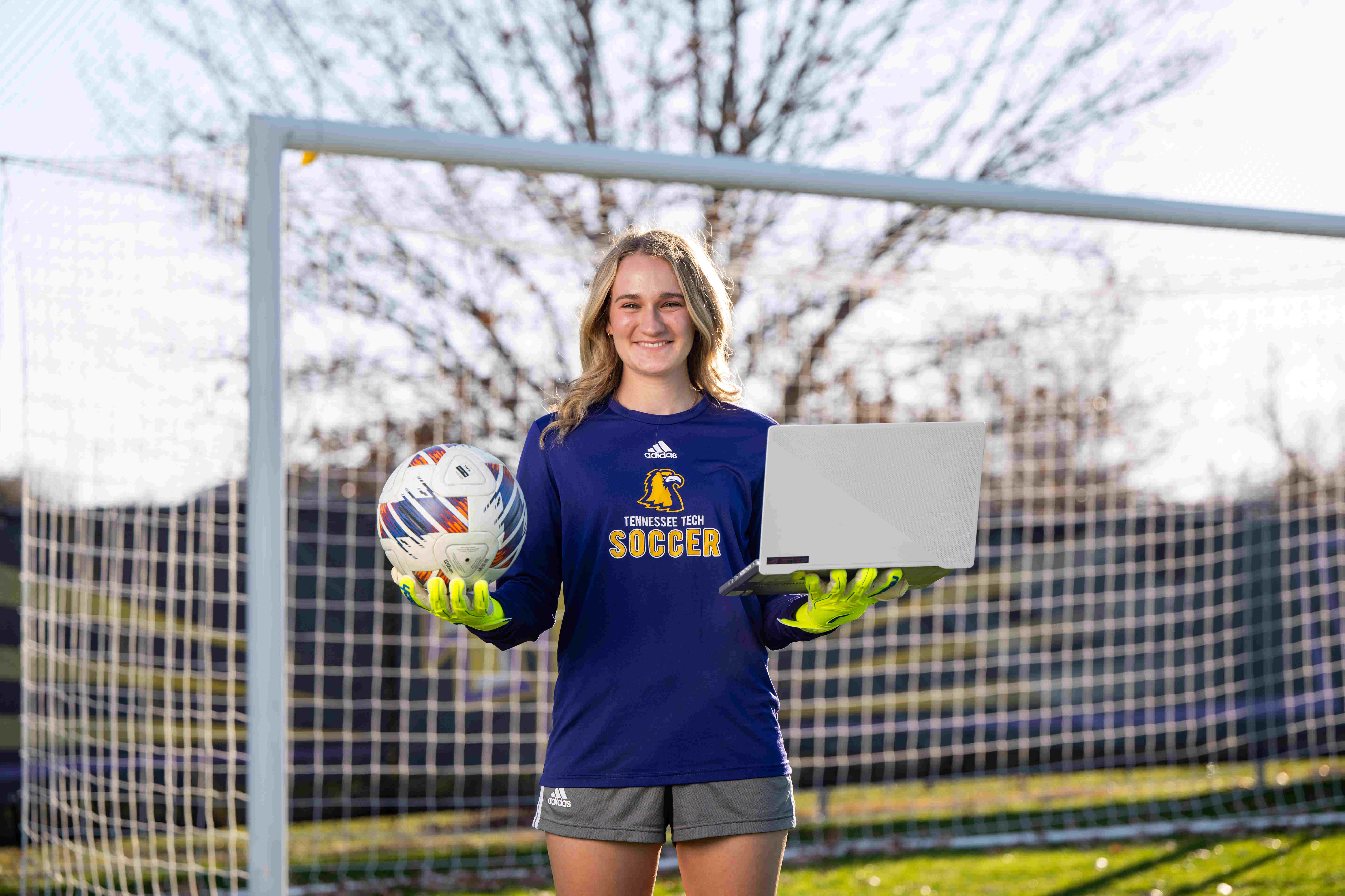 Addison Johnson on Tech's soccer field in front of the goal holding a soccer ball in one hand and her computer in the other