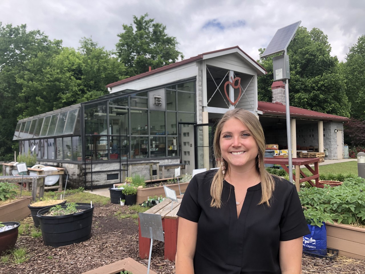 Abbey smiles at the camera. She is standing in front of a garden with a building in the background. The building has a greenhouse area and a sign on the front in the shape of an apple.