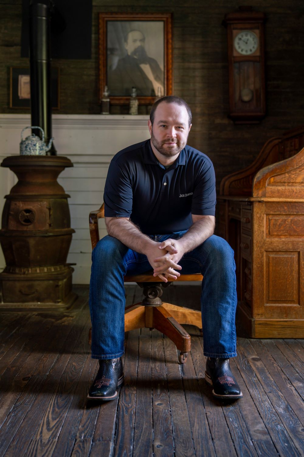 Chris Fletcher sits on a wooden office chair in front of an antique potbelly stove, an antique desk, and a portrait of Jack Daniels.