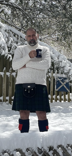Charles white is holding a mug and standing in the snow in a kilt.