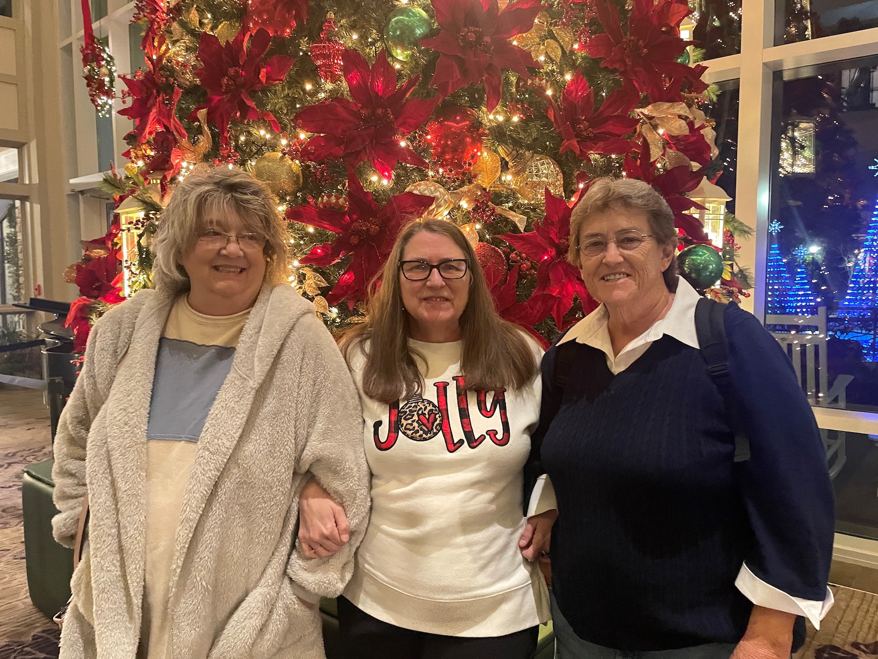 three women stand in front of a Christmas tree decorated in poinsettias