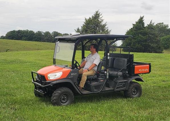A photo of a man riding a side-by-side work vehicle in a field.