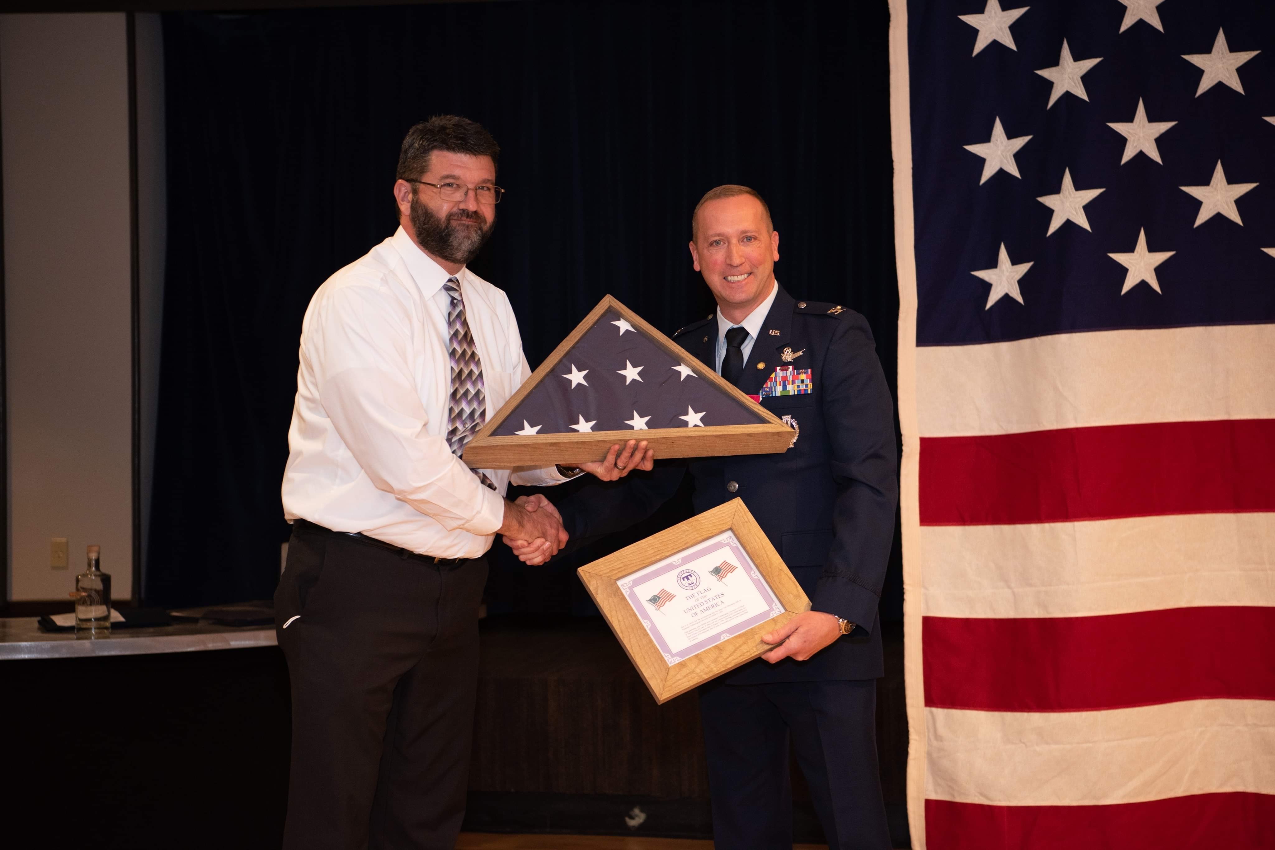 Two gentlemen are shaking hands while one is presented with the framed American Flag and a certificate. 