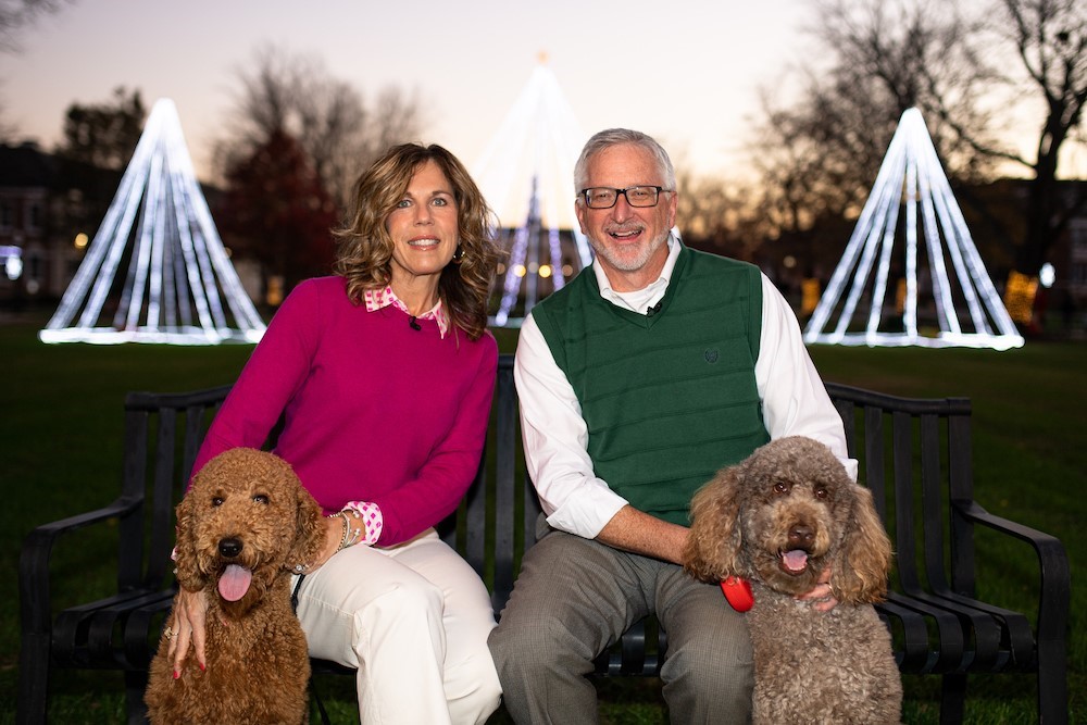 President Phil Oldham and First Lady Kari Oldham sit in front of the lights on the Quad at dusk with their two dogs.