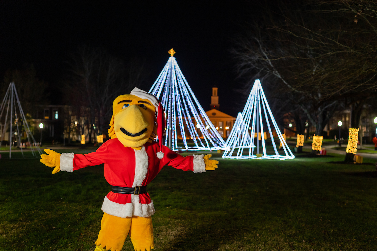 Awesome Eagle stands in front of the lit up quad. He is wearing a Santa hat and coat.