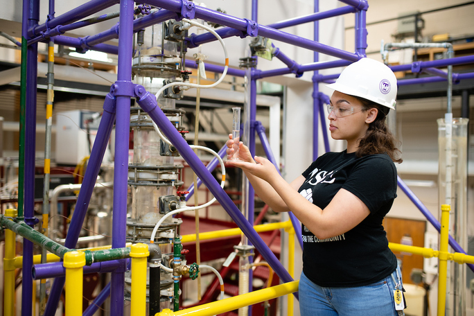 A student wearing a purple hard hat observes a clear liquid in a graduated cylinder.