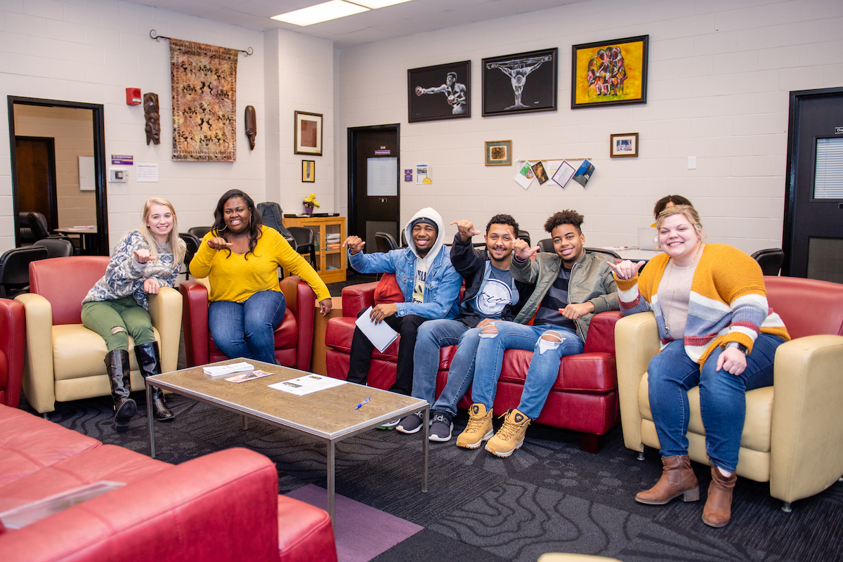A group of students sitting on couches in the BCC giving the wings up and smiling to the camera.