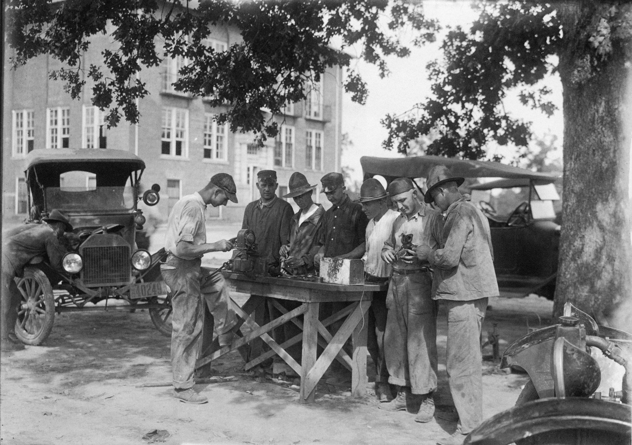 A black and white photo with a group of gentlemen standing around a workbench with 20s cars behind them.
