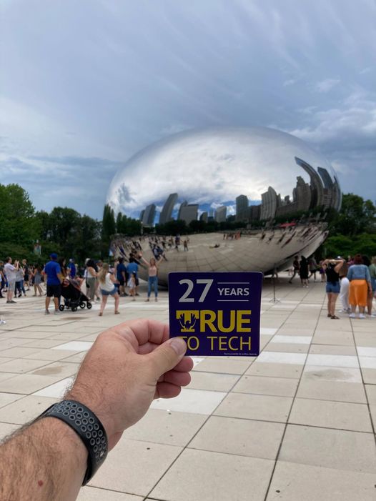a hand holding a 27 years True To Tech magnet in front of the Cloud Gate sculpture in Chicago