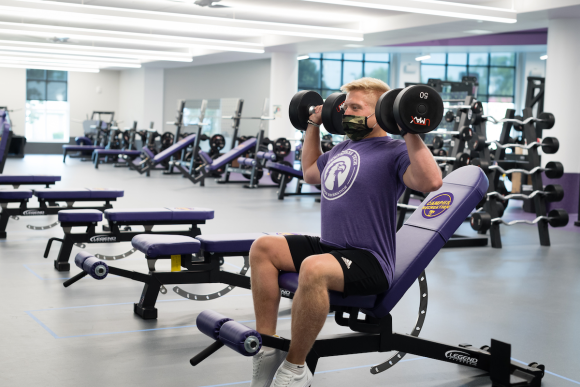 Photo of a student wearing a mask and using a purple weight bench and two dumbbells. 