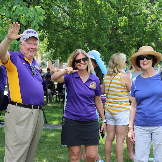 A photo from the 100th Anniversary Event. Dr. Bob Bell waves at the camera with Gloria Bell and Becky Magura. Becky is giving wings up.
