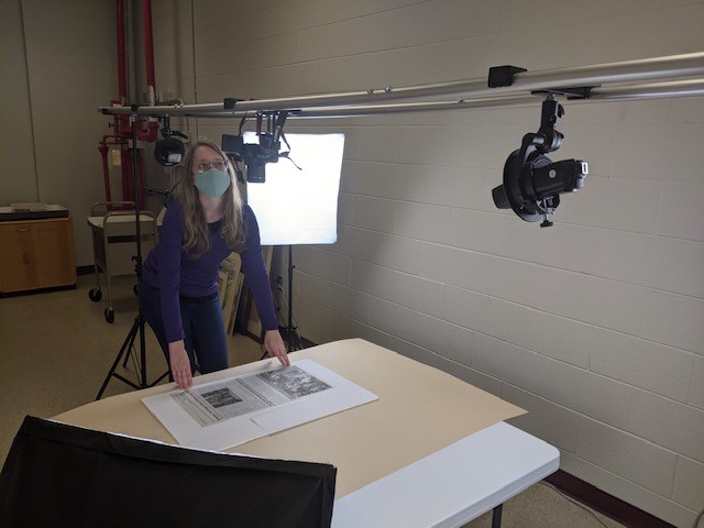 Assistant Archivist Hannah wears a mask and looks up at a camera on a rail system with a light box behind her. A large format print lays on the table in front of her.