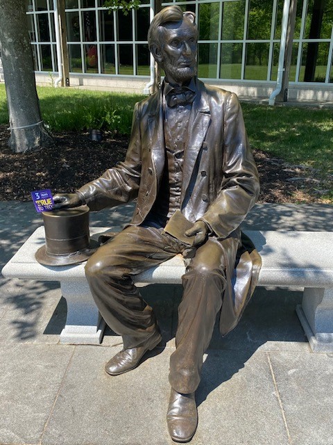 a bronze statue of Abraham Lincoln sitting on a concrete bench - there is a 3 years True To Tech magnet placed in his right hand, which lays atop a bronze stovepipe hat