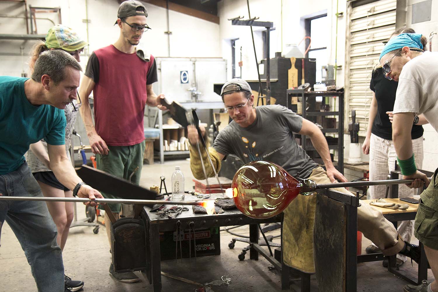 Several students observe three people working on a blown glass piece.
