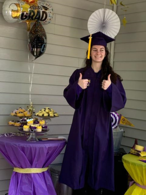 Photo of a graduate in purple regalia giving thumbs up. There is a table beside the graduate, covered in a purple table cloth, with trays of cupcakes and balloons. 